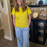 Woman in a yellow shirt and blue jeans standing in a room with a mirror and furniture.