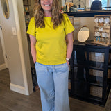 Woman in a yellow shirt and blue jeans standing in a room with a mirror and furniture.