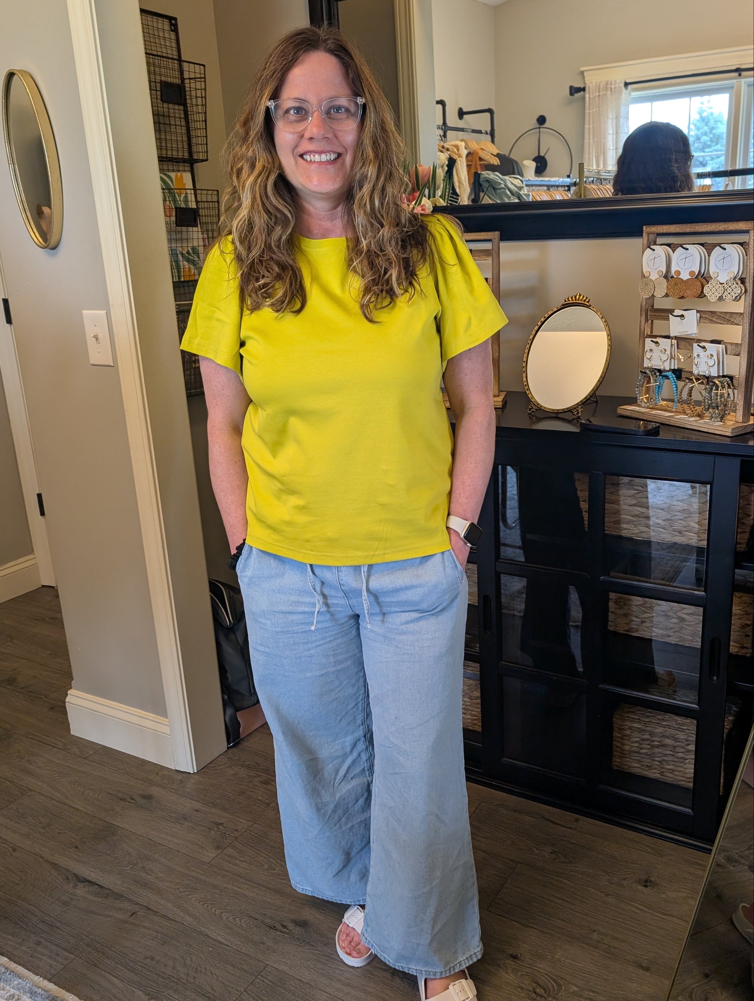 Woman in a yellow shirt and blue jeans standing in a room with a mirror and furniture.