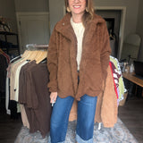 A woman wearing a brown teddy fleece jacket, blue jeans, and brown flats stands in a room with clothing racks and a patterned rug.