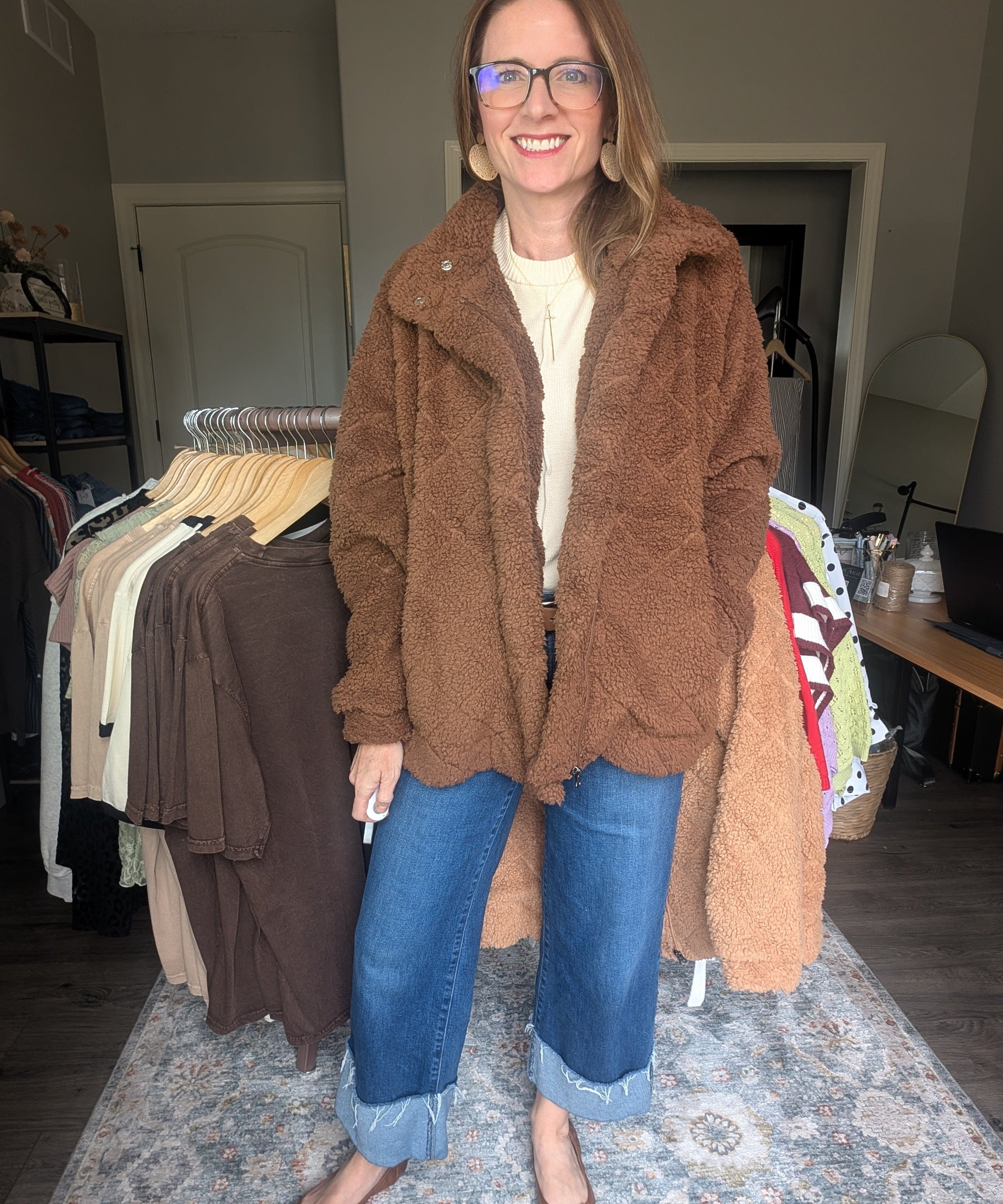 A woman wearing a brown teddy fleece jacket, blue jeans, and brown flats stands in a room with clothing racks and a patterned rug.