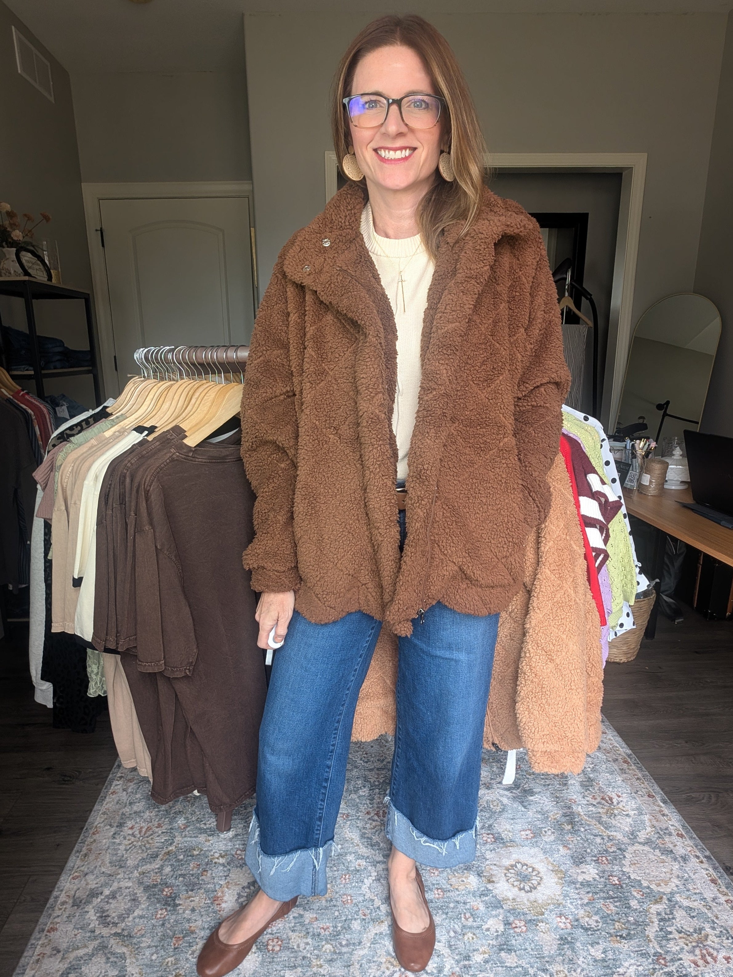 A woman wearing a brown teddy fleece jacket, blue jeans, and brown flats stands in a room with clothing racks and a patterned rug.