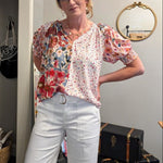 Woman wearing a floral blouse and white pants standing in a room with suitcases and a mirror.