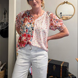 Woman wearing a floral blouse and white pants standing in a room with suitcases and a mirror.