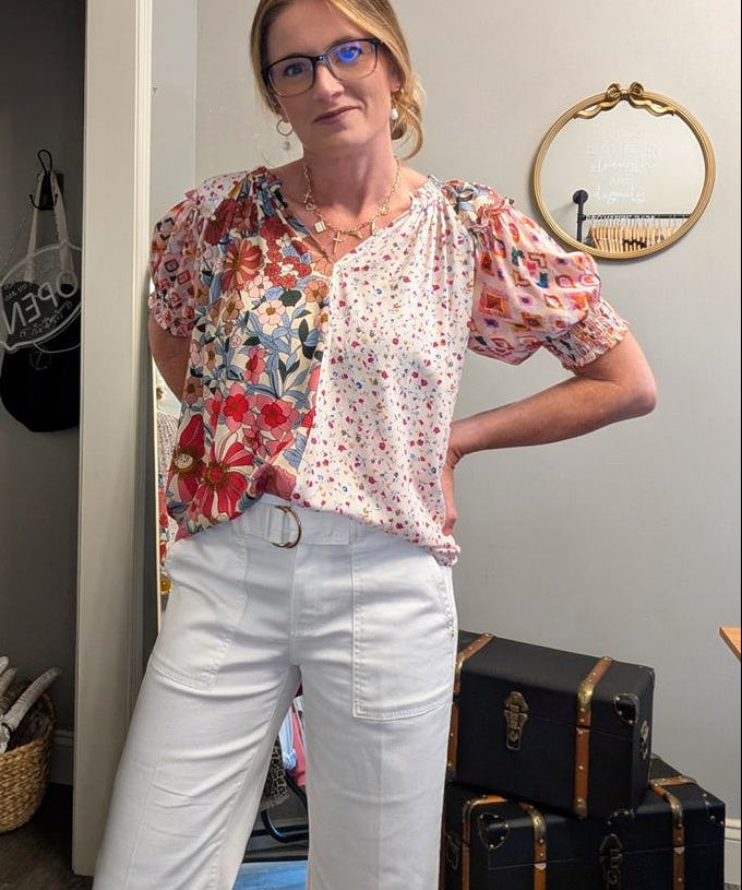 Woman wearing a floral blouse and white pants standing in a room with suitcases and a mirror.