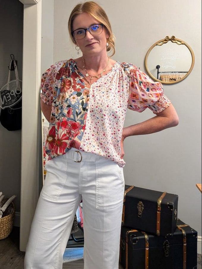 Woman wearing a floral blouse and white pants standing in a room with suitcases and a mirror.