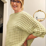 Woman wearing a light green sweater and white pants standing in a room with a television and decor items.