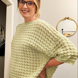 Woman wearing a light green sweater and white pants standing in a room with a television and decor items.