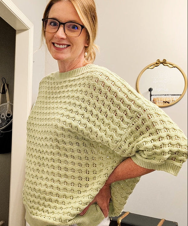 Woman wearing a light green sweater and white pants standing in a room with a television and decor items.