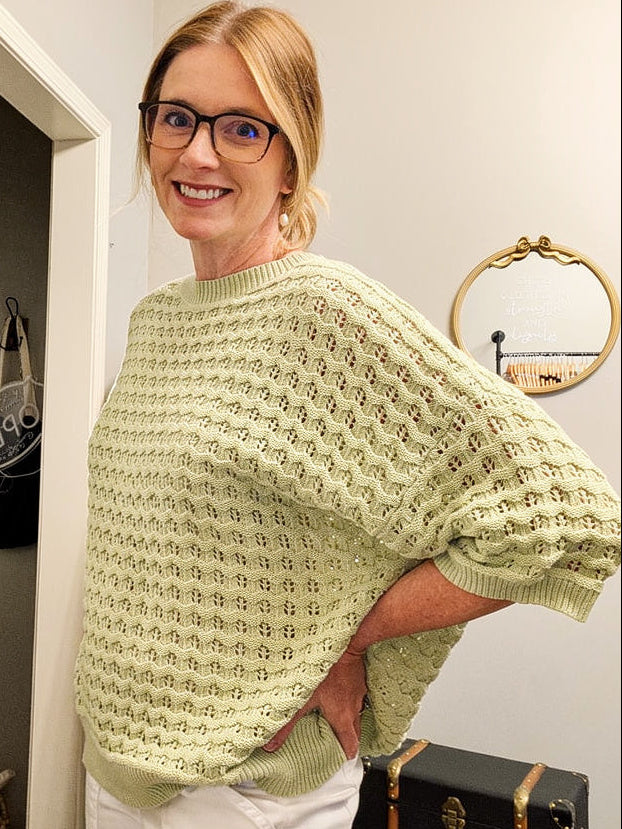 Woman wearing a light green sweater and white pants standing in a room with a television and decor items.