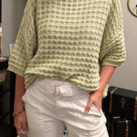 Woman wearing a light green sweater and white pants indoors.