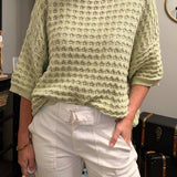 Woman wearing a light green sweater and white pants indoors.