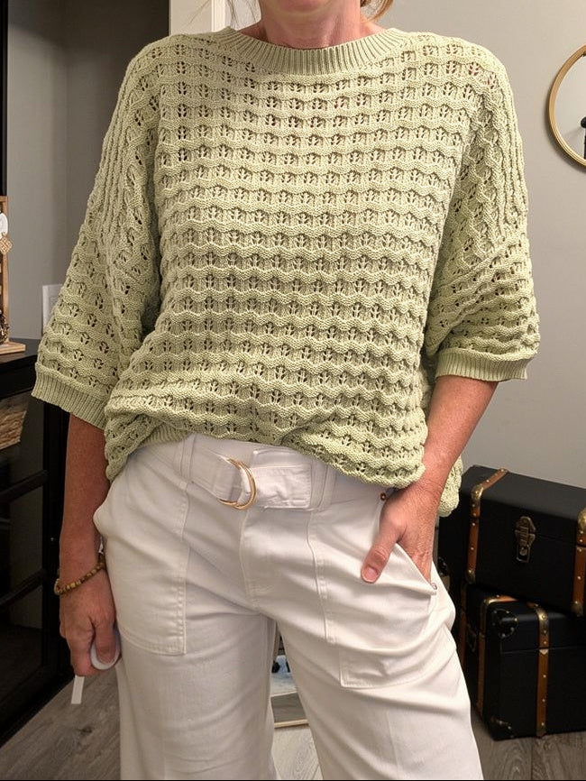 Woman wearing a light green sweater and white pants indoors.