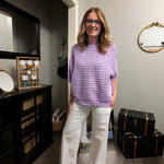 Woman in a purple sweater and white pants standing in a room with furniture and decor.
