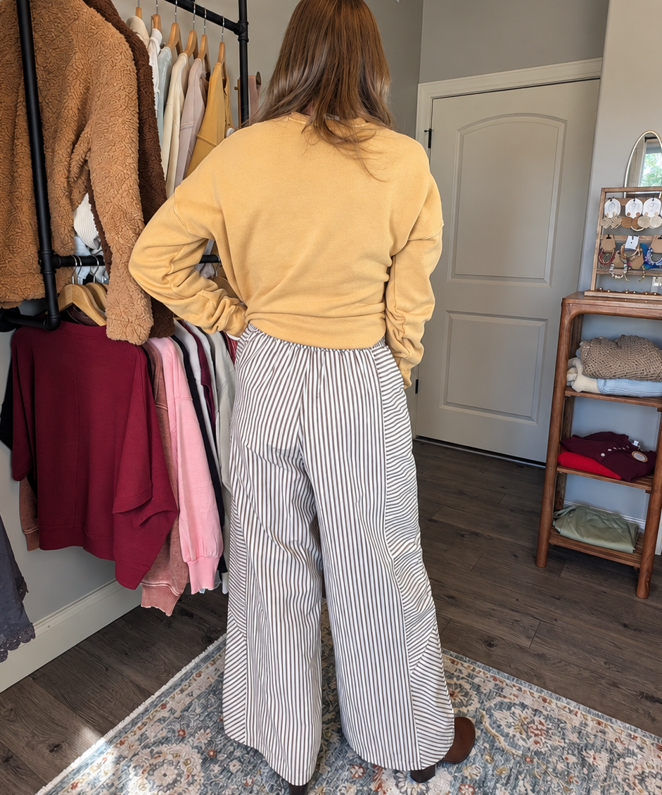 A woman stands with her back to the camera in a home closet environment, wearing a mustard yellow long-sleeve top and high-waisted, wide-leg gray and white striped trousers.