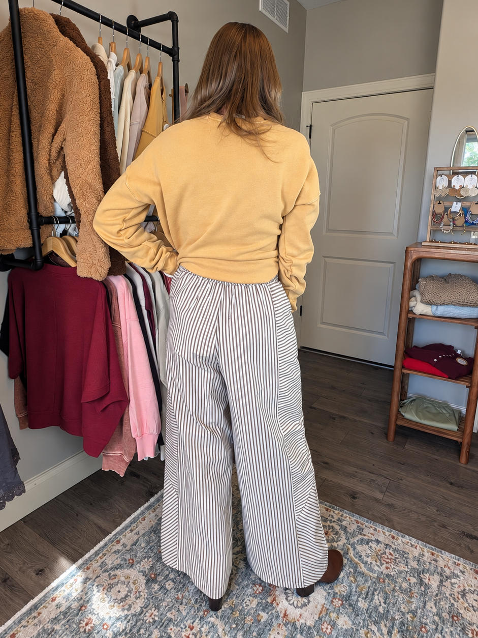 A woman stands with her back to the camera in a home closet environment, wearing a mustard yellow long-sleeve top and high-waisted, wide-leg gray and white striped trousers.