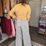 A woman stands with her back to the camera in a home closet environment, wearing a mustard yellow long-sleeve top and high-waisted, wide-leg gray and white striped trousers.