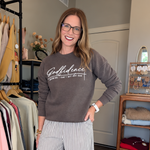 A woman wearing a brown crewneck sweatshirt with Godfidence printed on the front and striped wide-leg pants, standing in a room with clothing racks and a wooden floor.