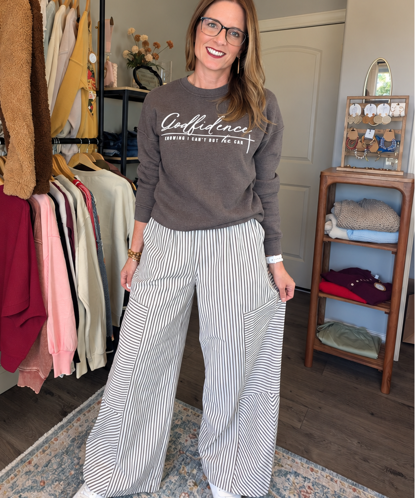 A woman stands in a room wearing a brown long-sleeve graphic sweatshirt and white-and-gray vertically striped wide-leg pants. Clothing racks and a wooden shelf are visible in the background.