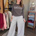 A woman stands in a room wearing a brown long-sleeve graphic sweatshirt and white-and-gray vertically striped wide-leg pants. Clothing racks and a wooden shelf are visible in the background.